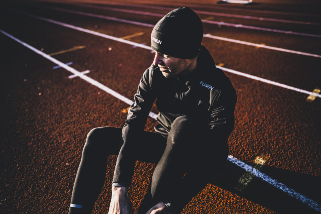 Man sitting tieing his shoes on running track wearing FUSION Sportswar jacket and beanie